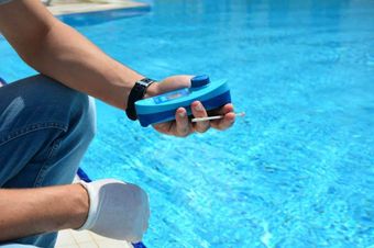Person holding a blue pool testing device beside a swimming pool