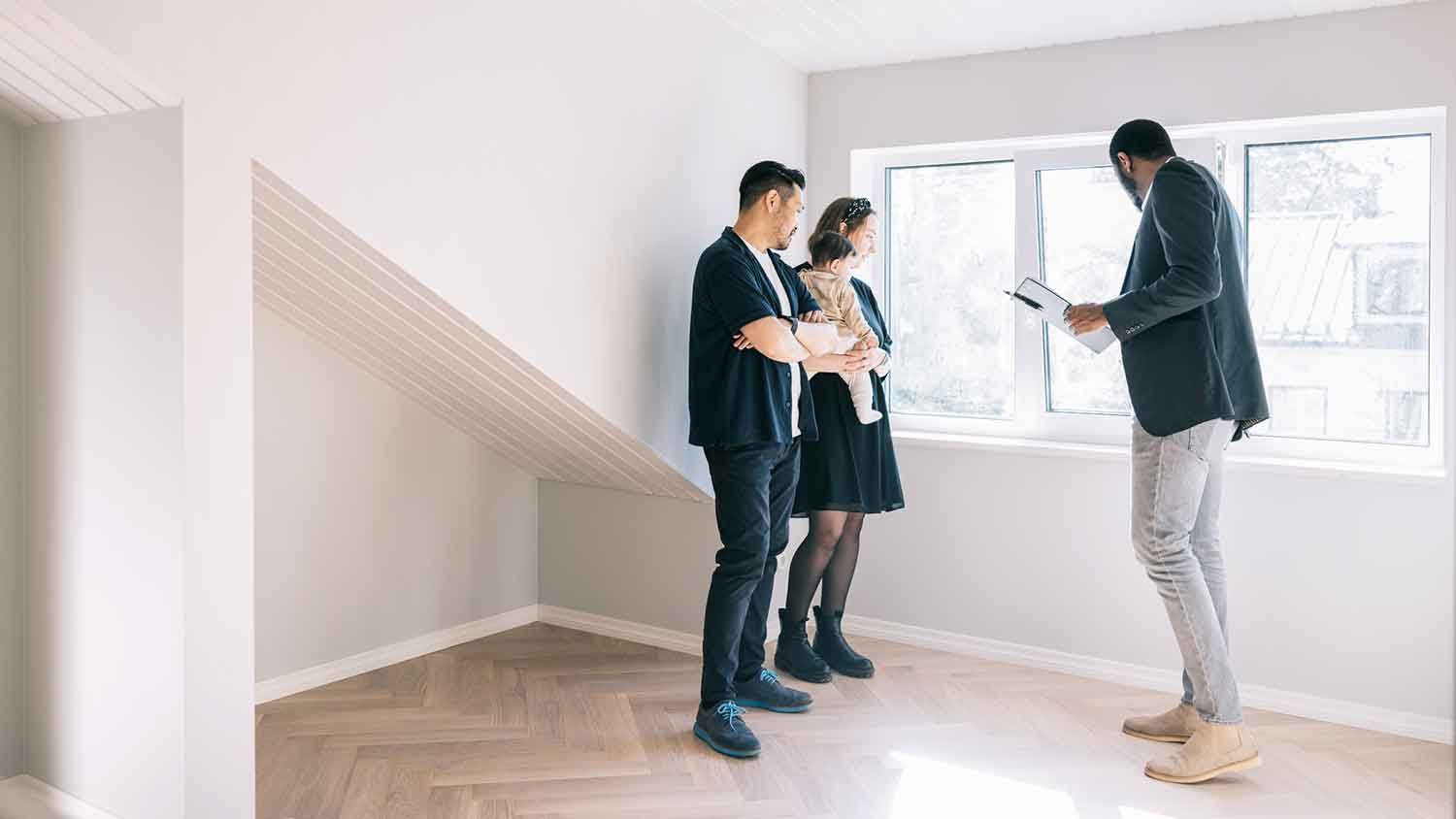 Three people in a bright empty room discuss a house while one gestures near a window