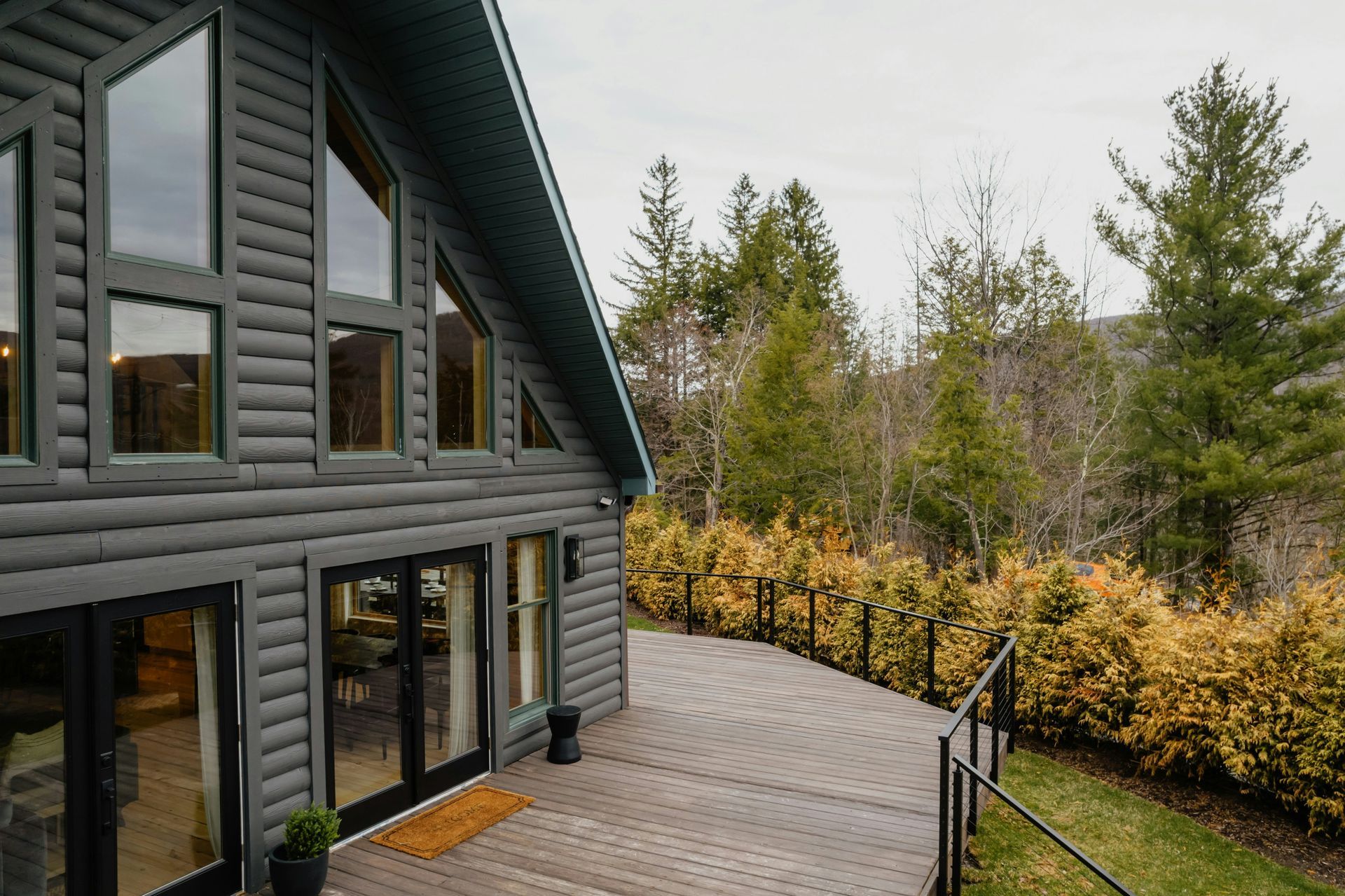 Dark gray log cabin with large windows and a wooden deck beside trees and a hedge
