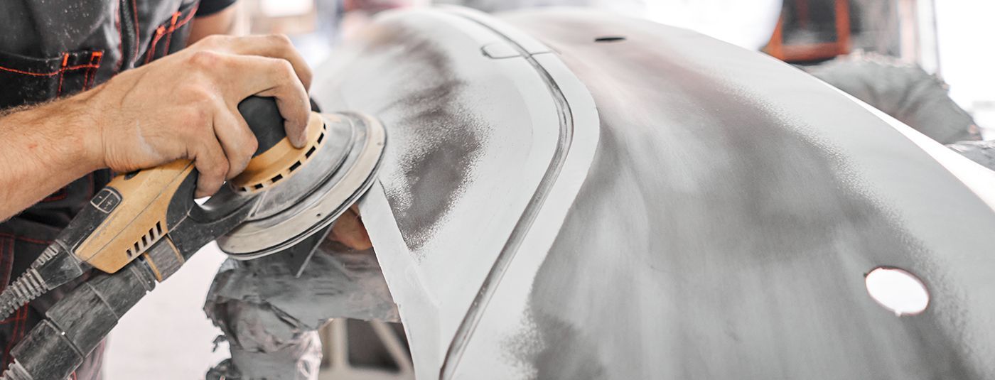 A person uses a power sander to smooth a primed car bumper in an automotive repair shop.