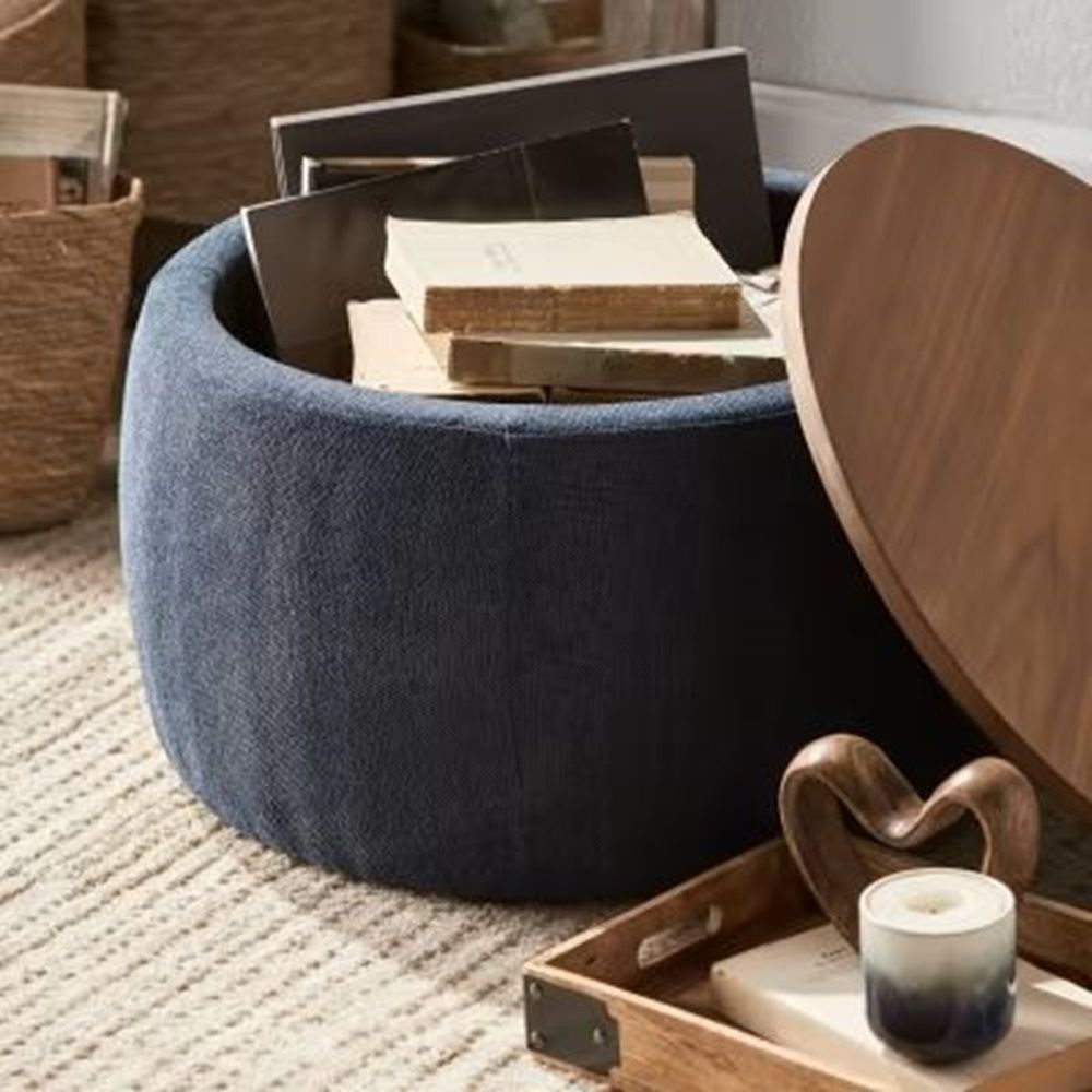 Un pouf contenitore rotondo blu navy con coperchio in legno, pieno di libri. È appoggiato su un tappeto color crema, con un vassoio e una scultura a forma di cuore nelle vicinanze.