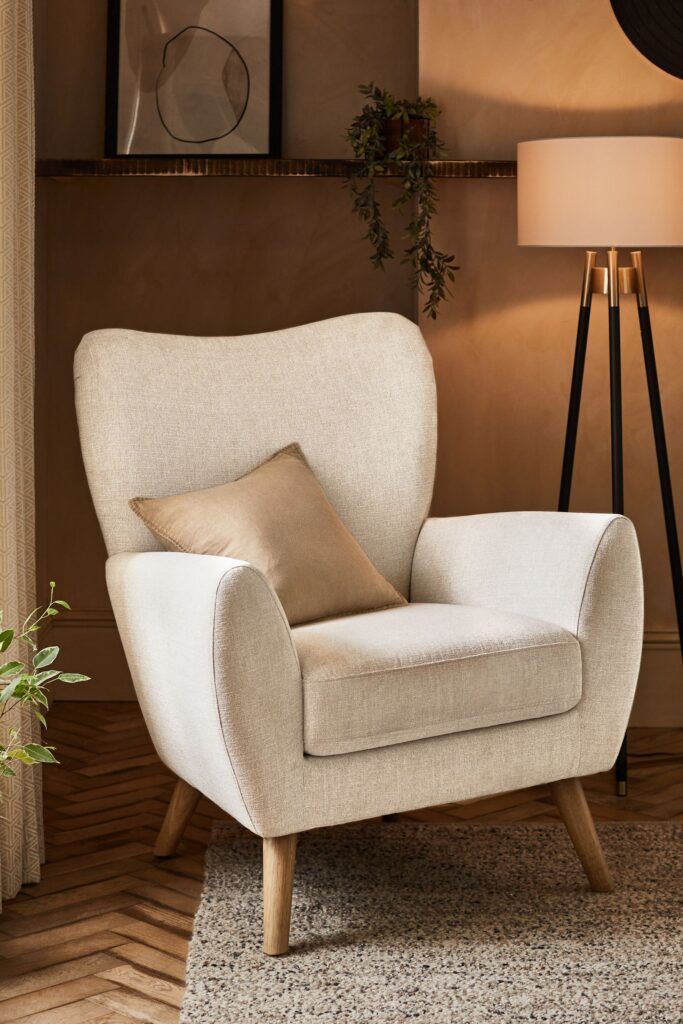 Poltrona color crema con cuscino beige in un accogliente soggiorno. Sullo sfondo, una lampada da terra e una mensola decorativa.