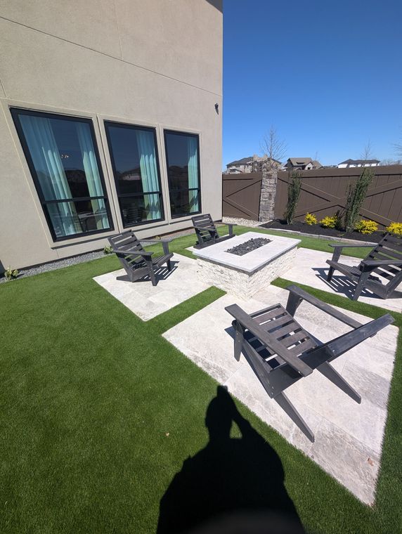 Patio with black lounge chairs on artificial grass beside a beige building and windows, under a bright blue sky