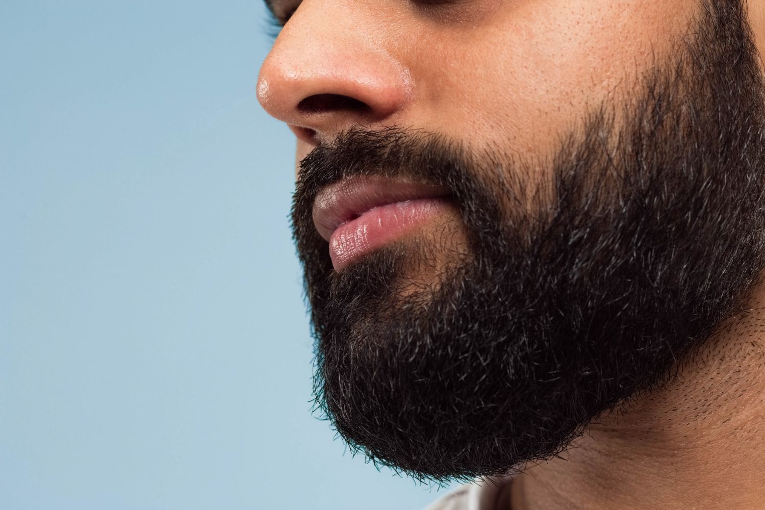 Close-up do rosto de uma pessoa, com foco na barba escura e parte do nariz e dos lábios, contra um fundo azul claro.