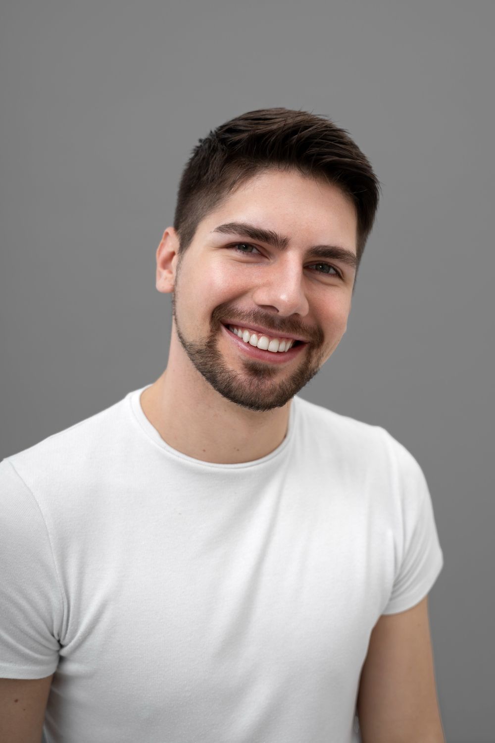 Homem com cabelos e barba escuros sorrindo, vestindo uma camiseta branca, sobre um fundo cinza.