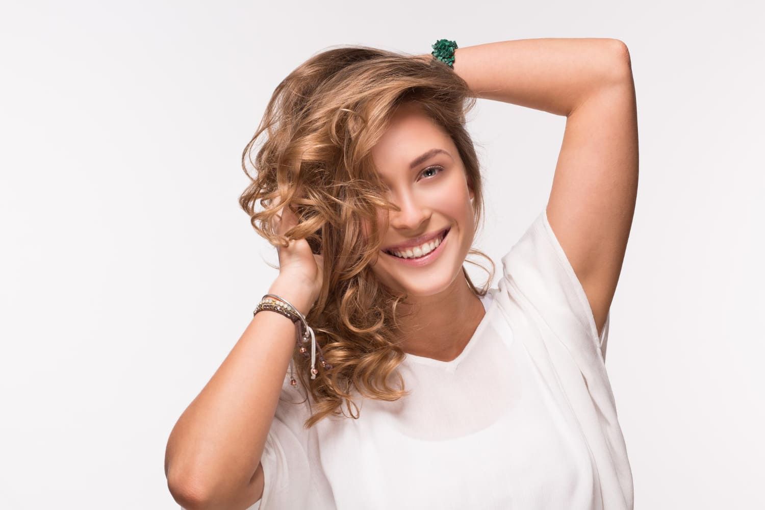 Mulher com cabelos castanhos cacheados sorri, braços levantados, vestindo uma blusa branca, posando contra um fundo branco.