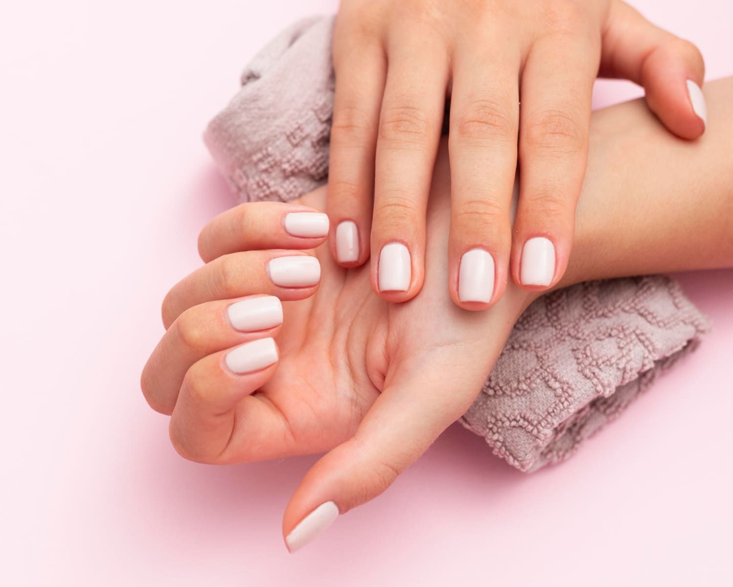 Mãos com unhas rosa-claro apoiadas sobre uma toalha cinza dobrada, tudo sobre um fundo rosa.