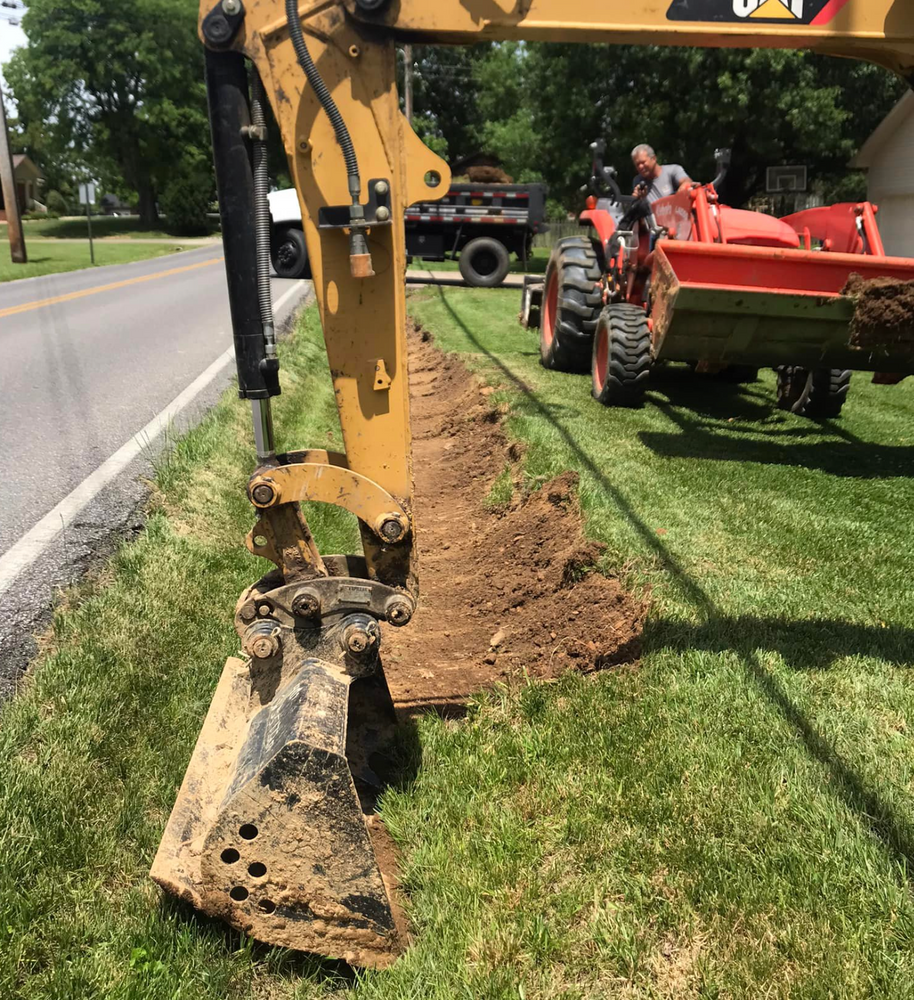 Excavator digging a trench beside a road, with a tractor and trailer nearby.