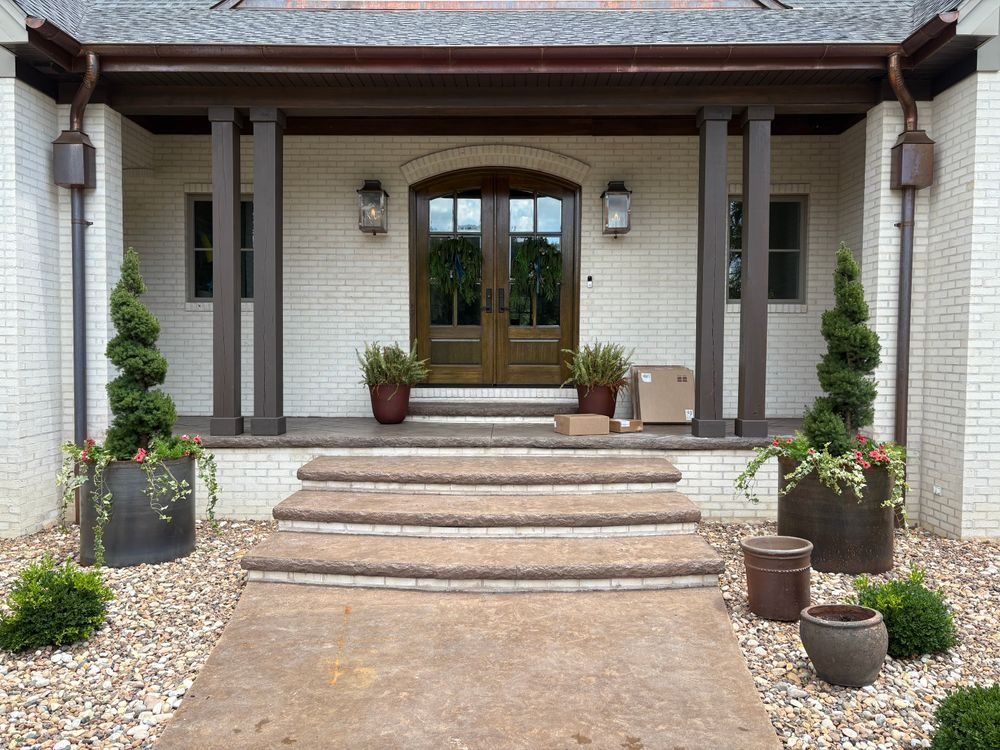 Front porch with steps, door, plants, and package on concrete walkway. White brick, brown trim.