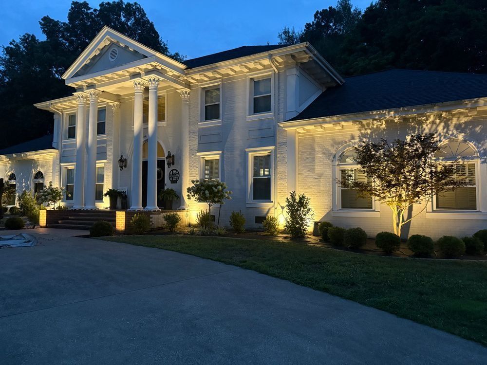 White mansion at dusk, lit with golden lights. Tall columns, dark roof, and driveway.