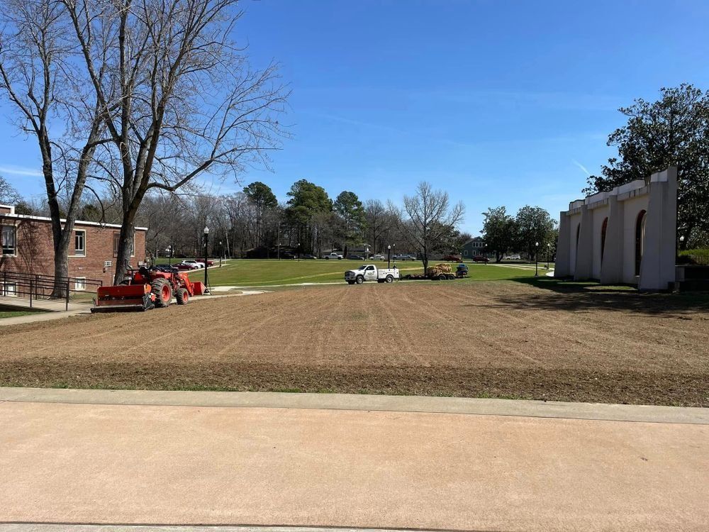 Machines preparing a grassy area on a sunny day, with buildings and trees in the background.