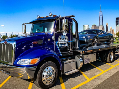 Blue tow truck carrying a black car in a city parking lot with skyline in the background