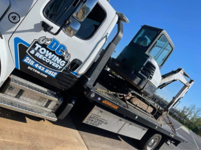 Tow truck hauling a black excavator on a flatbed in a sunny roadside setting