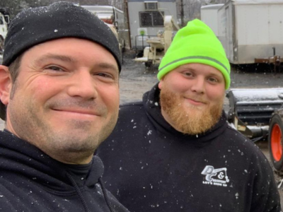 Two people smiling outdoors in winter hats, with snow falling and trucks in the background