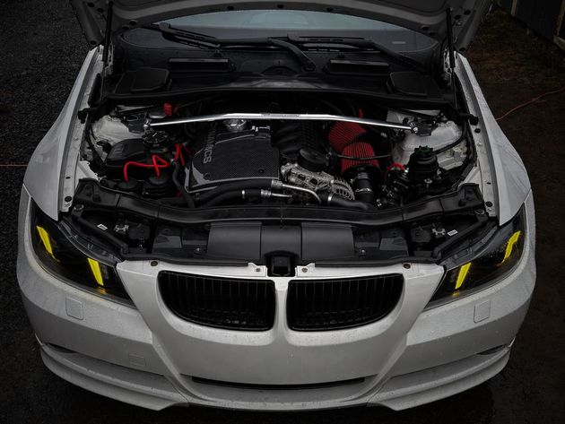 Open BMW engine bay with hood raised, showing red engine components and headlights on a dark background