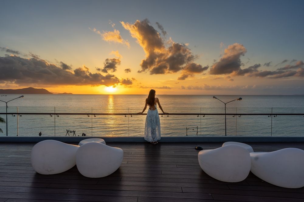 A woman in a white dress is standing on a balcony overlooking the ocean at sunset.