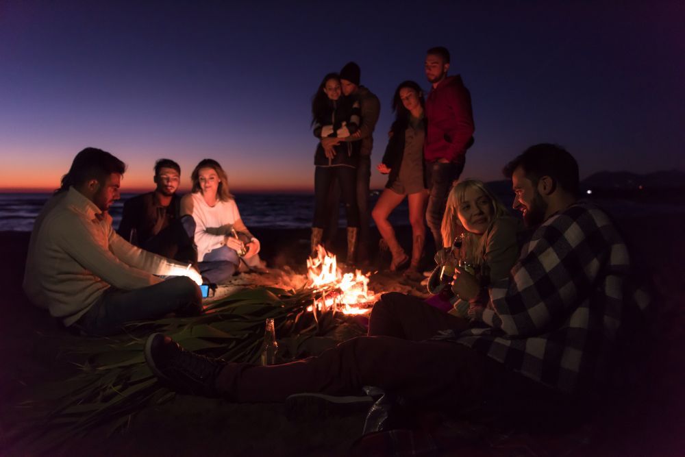 A group of people are sitting around a campfire on the beach at night.