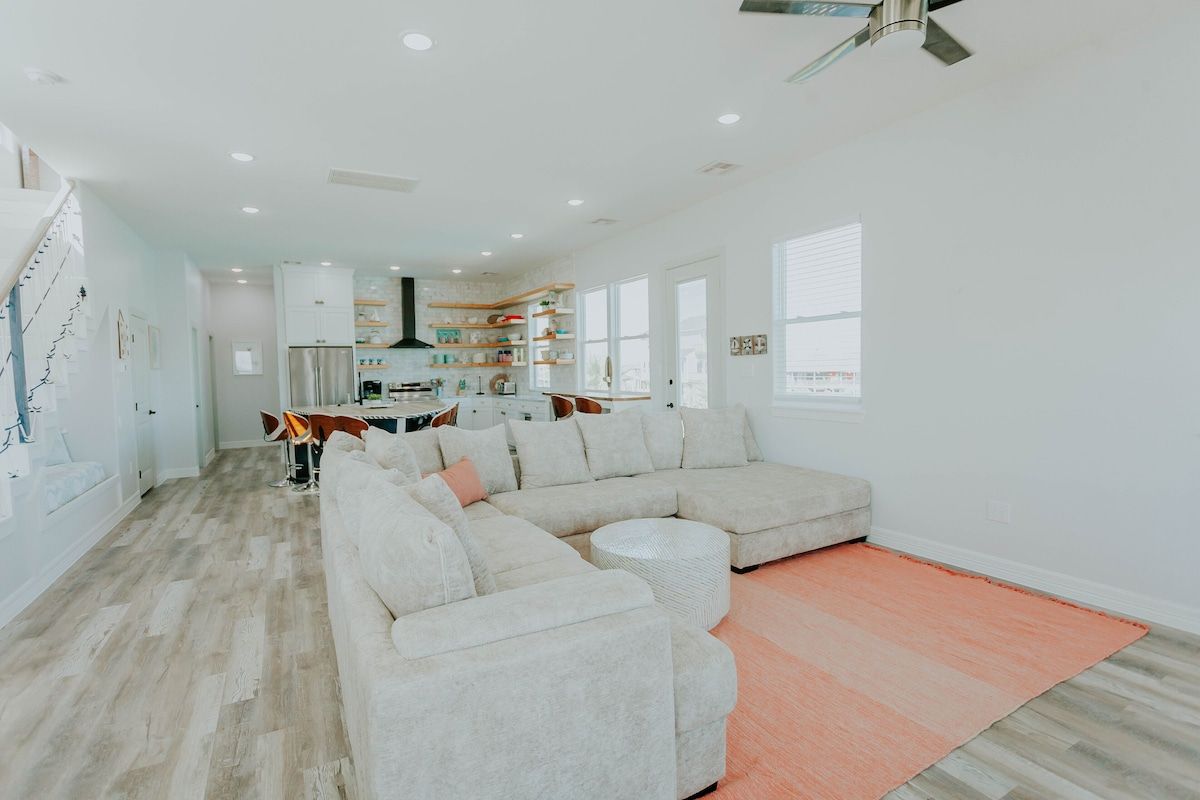 A spacious, bright open-plan living room with a large white sectional sofa on a peach rug, facing a kitchen in the back.