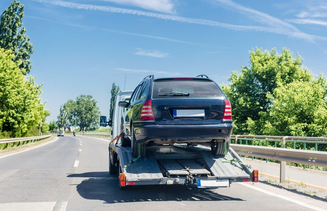 Black SUV on a tow truck driving on a sunny road with trees and guardrails