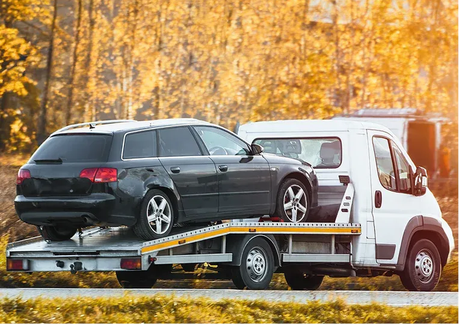 Tow truck carrying a black wagon and a white van on a road with autumn trees behind them
