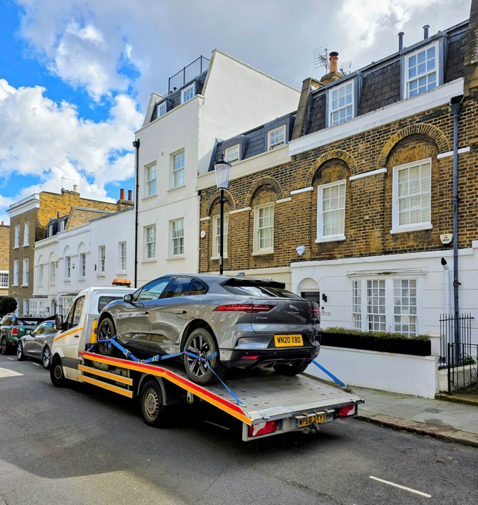 Grey car on a tow truck in a residential street with white and brick houses under a blue sky