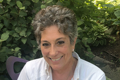 A woman in a white shirt is smiling in front of a bush.