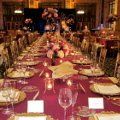 A long table with plates , glasses , and flowers on it in a restaurant.