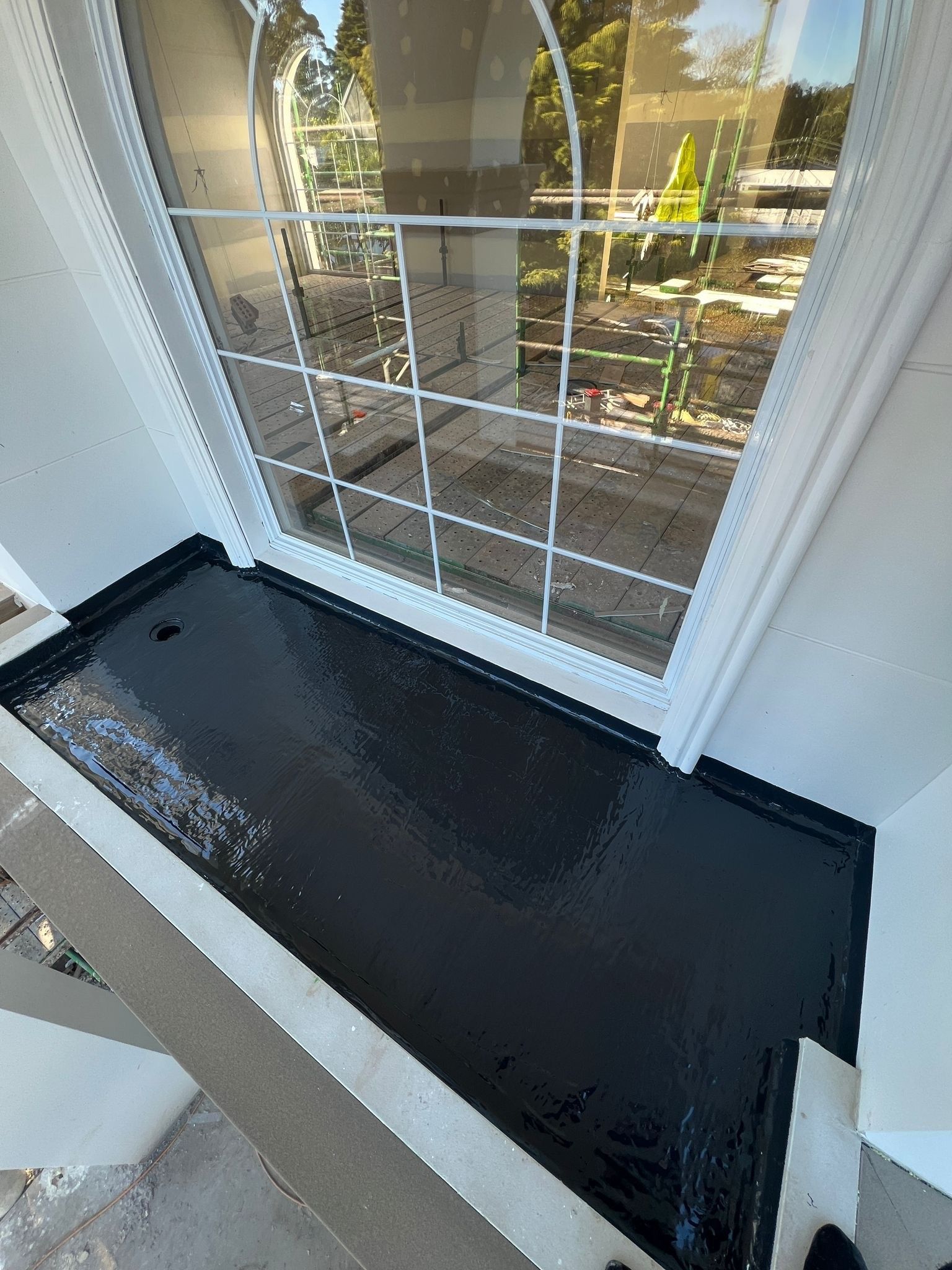 Small black balcony with white walls and a drain hole beside a glass door, overlooking a garden Small black balcony with white walls and a drain hole beside a glass door, overlooking a garden