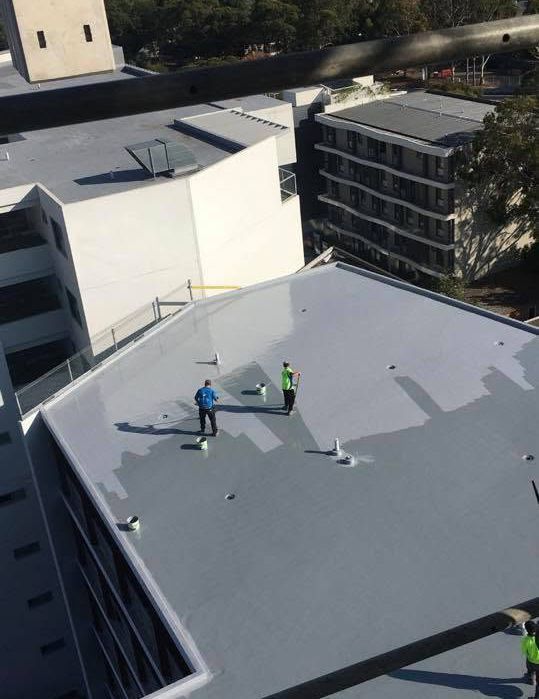 Two workers on a gray rooftop, one in blue and one in neon green, inspecting the roof.
