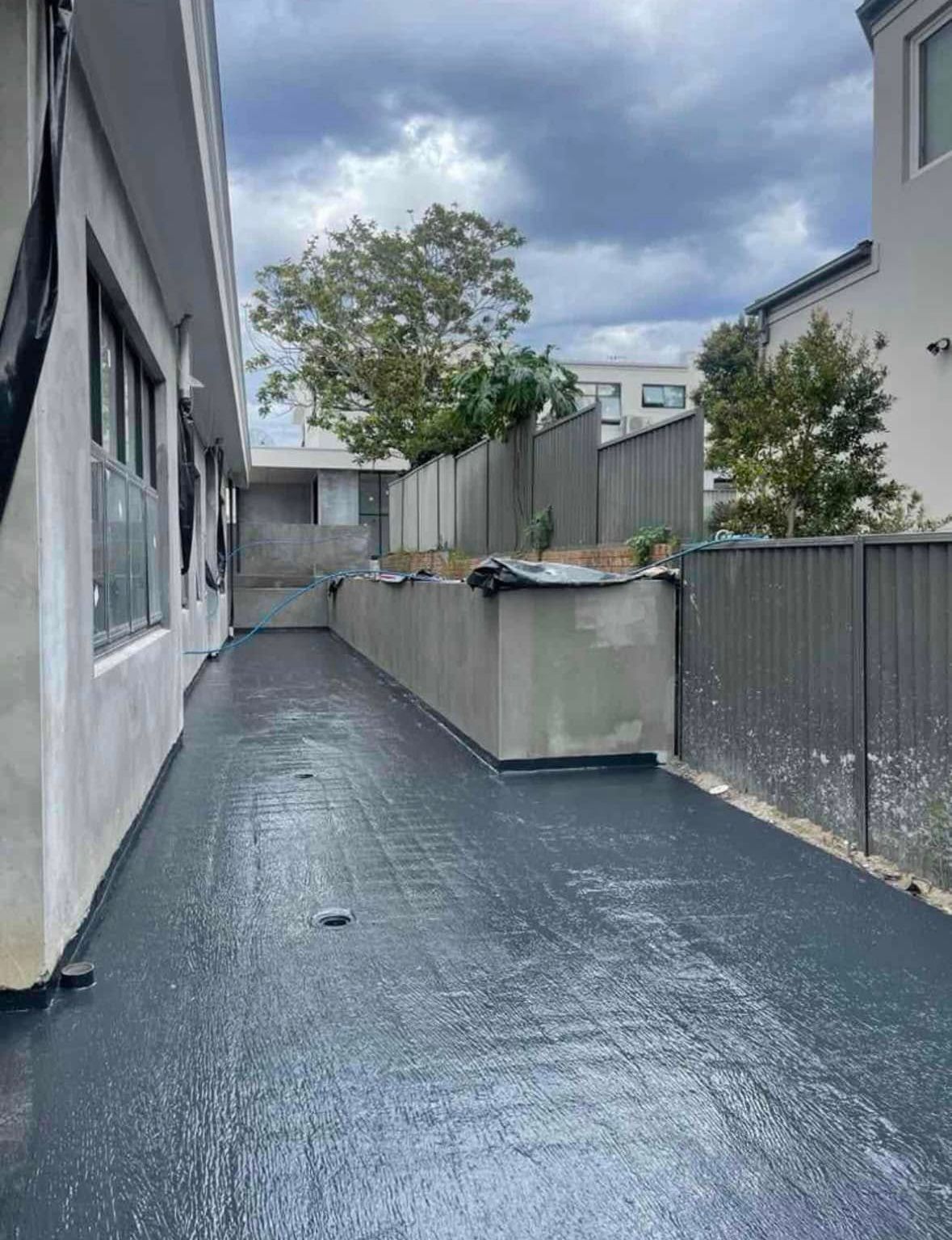 Narrow outdoor walkway beside buildings, with gray paving, fences, and potted plants under a cloudy sky Narrow outdoor walkway beside buildings, with gray paving, fences, and potted plants under a cloudy sky