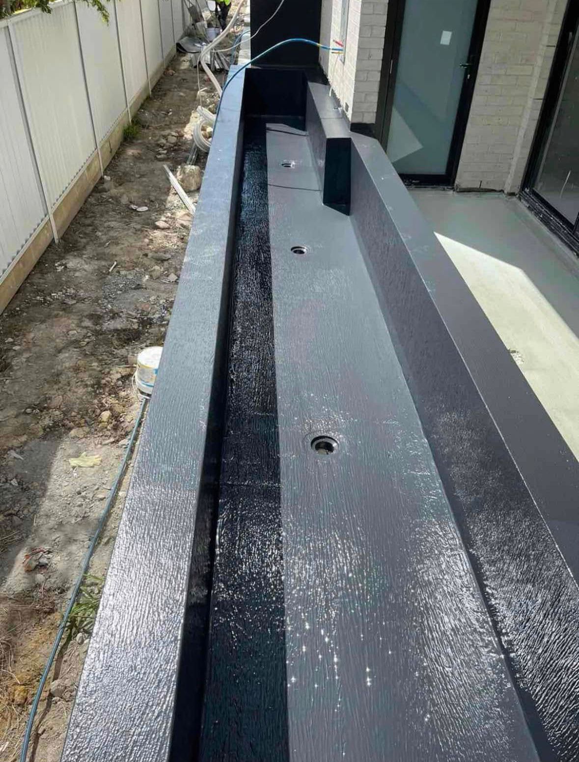 Black metal drainage channel beside a building, with several circular drain openings and a gravel edge. Black metal drainage channel beside a building, with several circular drain openings and a gravel edge.