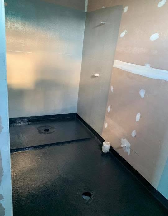 Unfinished bathroom with black waterproof floor, white walls, and patchy drywall repairs.