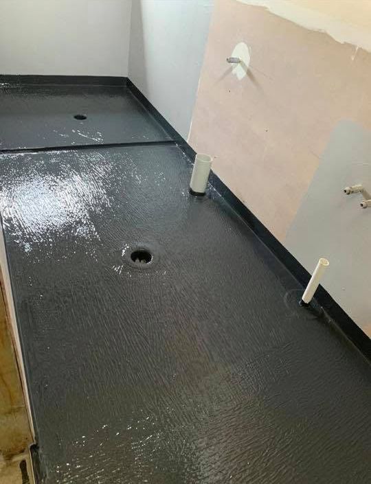 Freshly coated black bathroom floor with drain holes and exposed plumbing stubs against unfinished walls