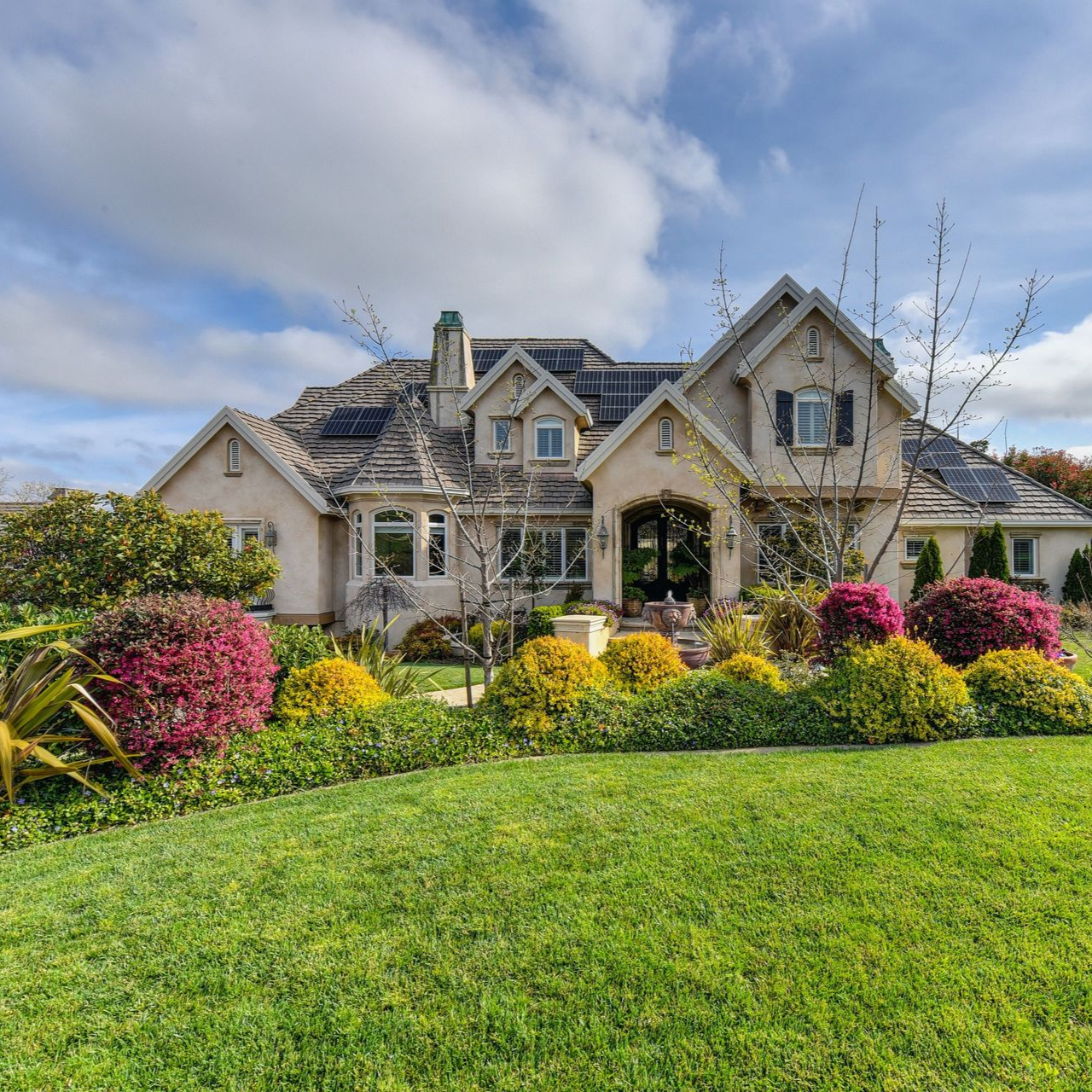 Large stone house with a gray roof, colorful flower beds, and a green lawn under a cloudy sky