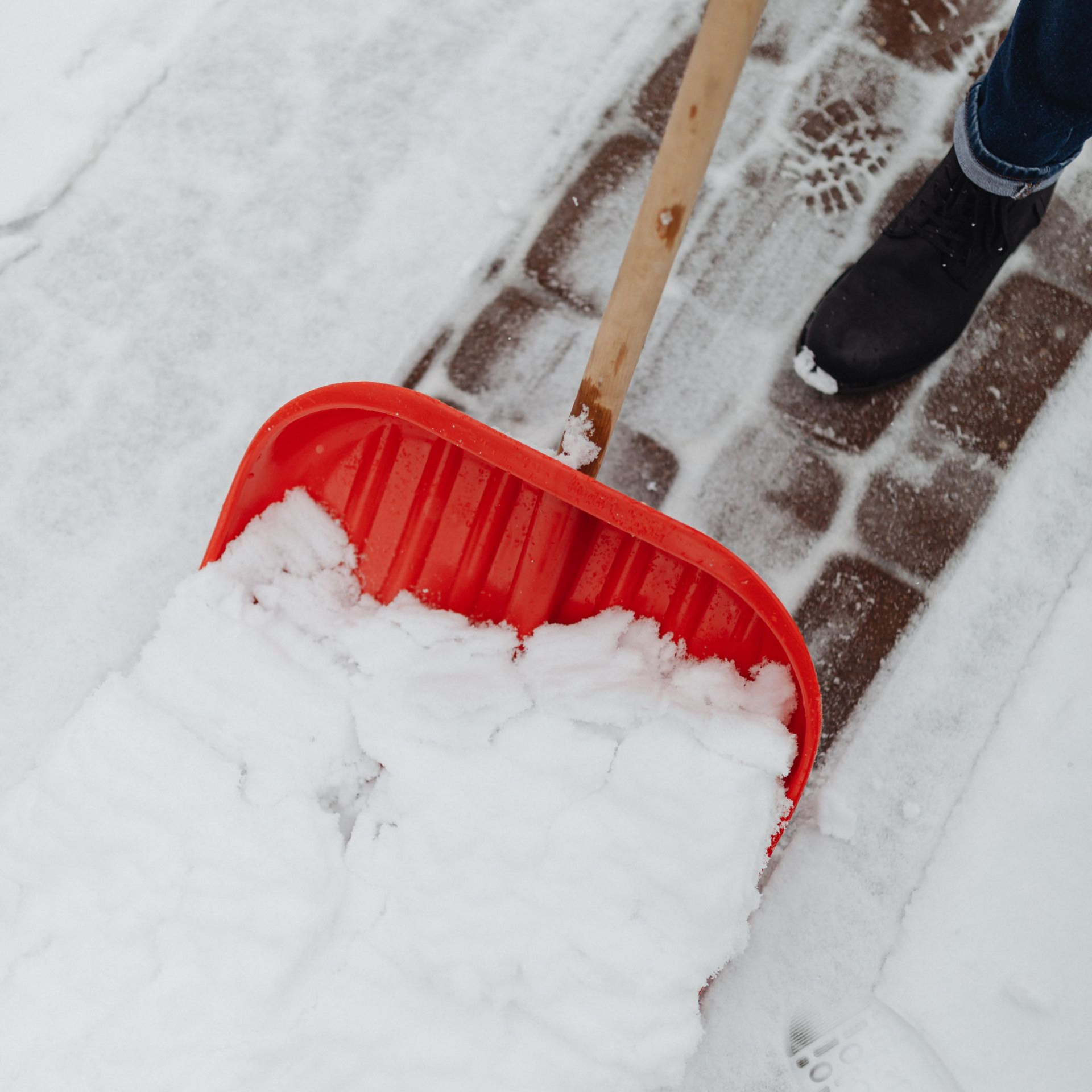 Red snow shovel full of snow on a snowy wooden deck, with a person’s boot nearby