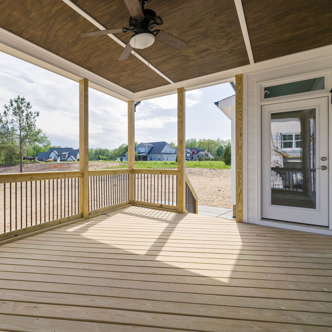 Covered porch with wood plank floor, railing, ceiling fan, and a white door overlooking a grassy yard