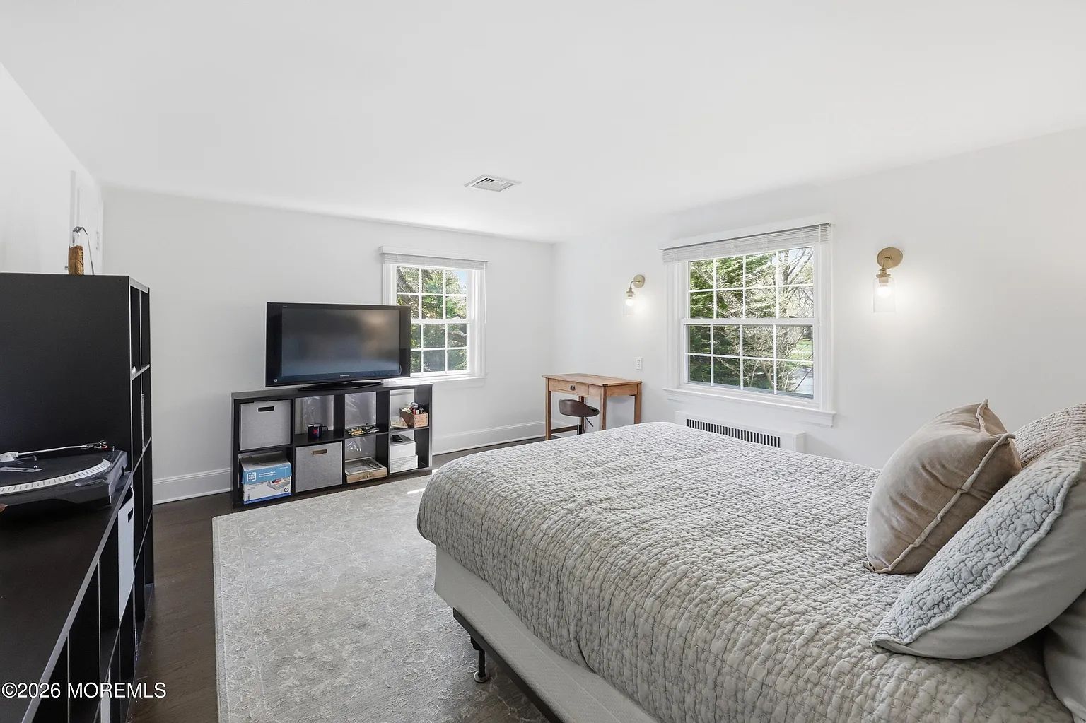 Bright bedroom with a large bed, TV stand, and two windows letting in natural light.