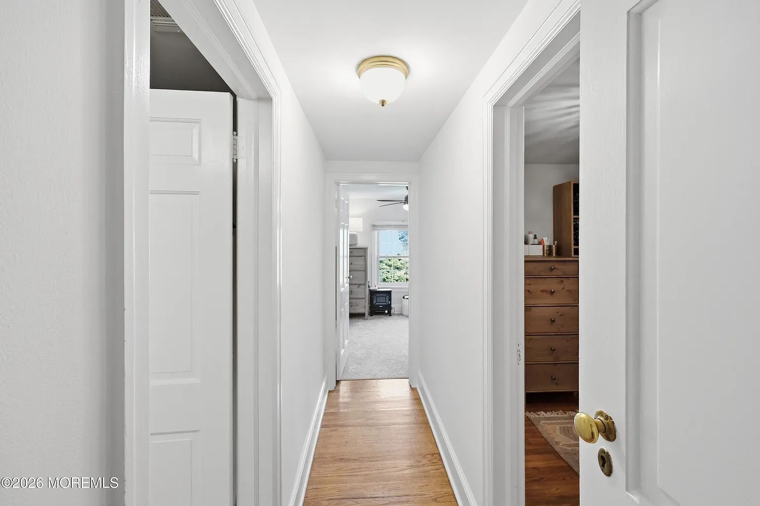 Narrow white hallway with wood floor, open doors, and a glimpse of a bright room at the end.