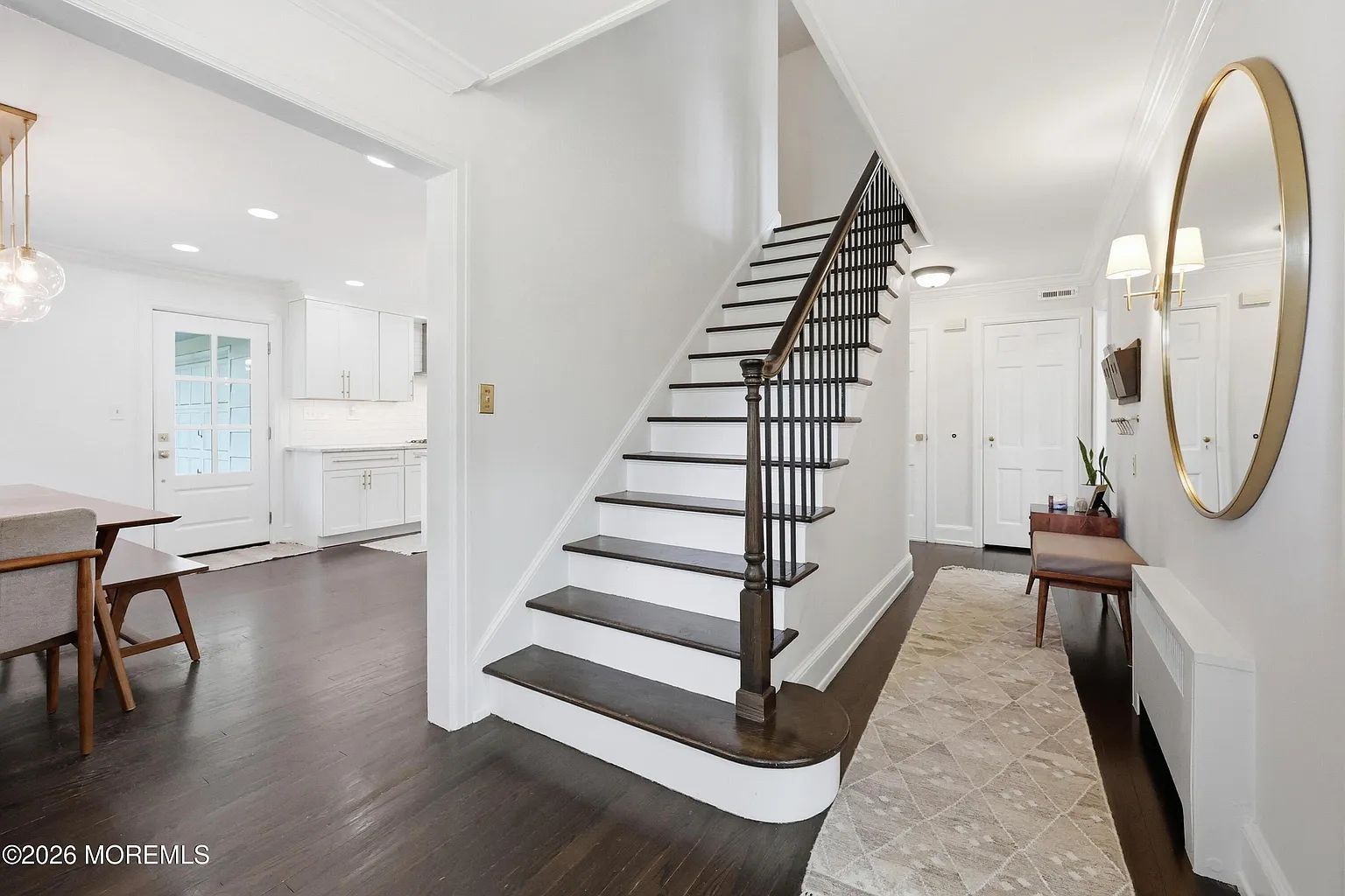 Bright hallway with black staircase, white walls, dark wood floors, and kitchen visible to the left.