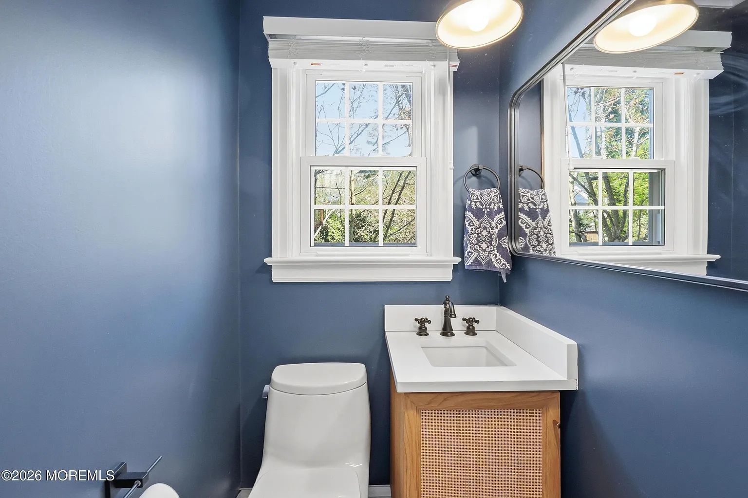 Blue bathroom with white sink, toilet, mirror, and two windows above a wicker cabinet.