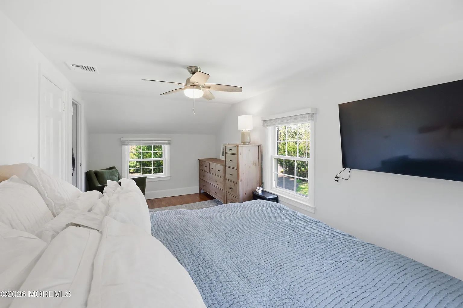 Bright white bedroom with a blue patterned bedspread, wall-mounted TV, and windows letting in daylight