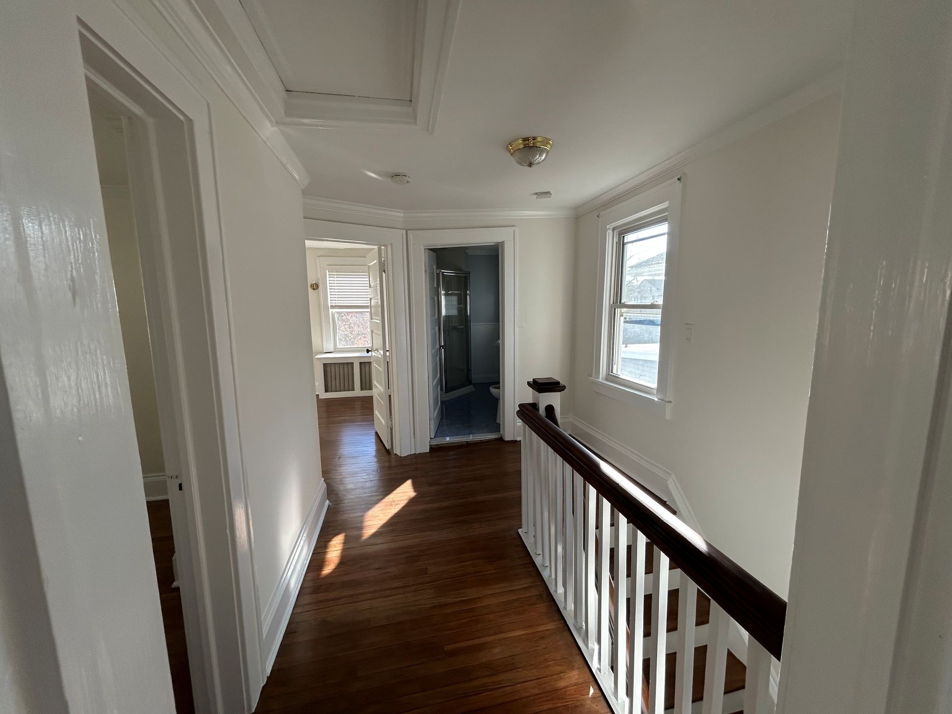 Narrow white hallway with dark wood floors, railing, and sunlight from windows.
