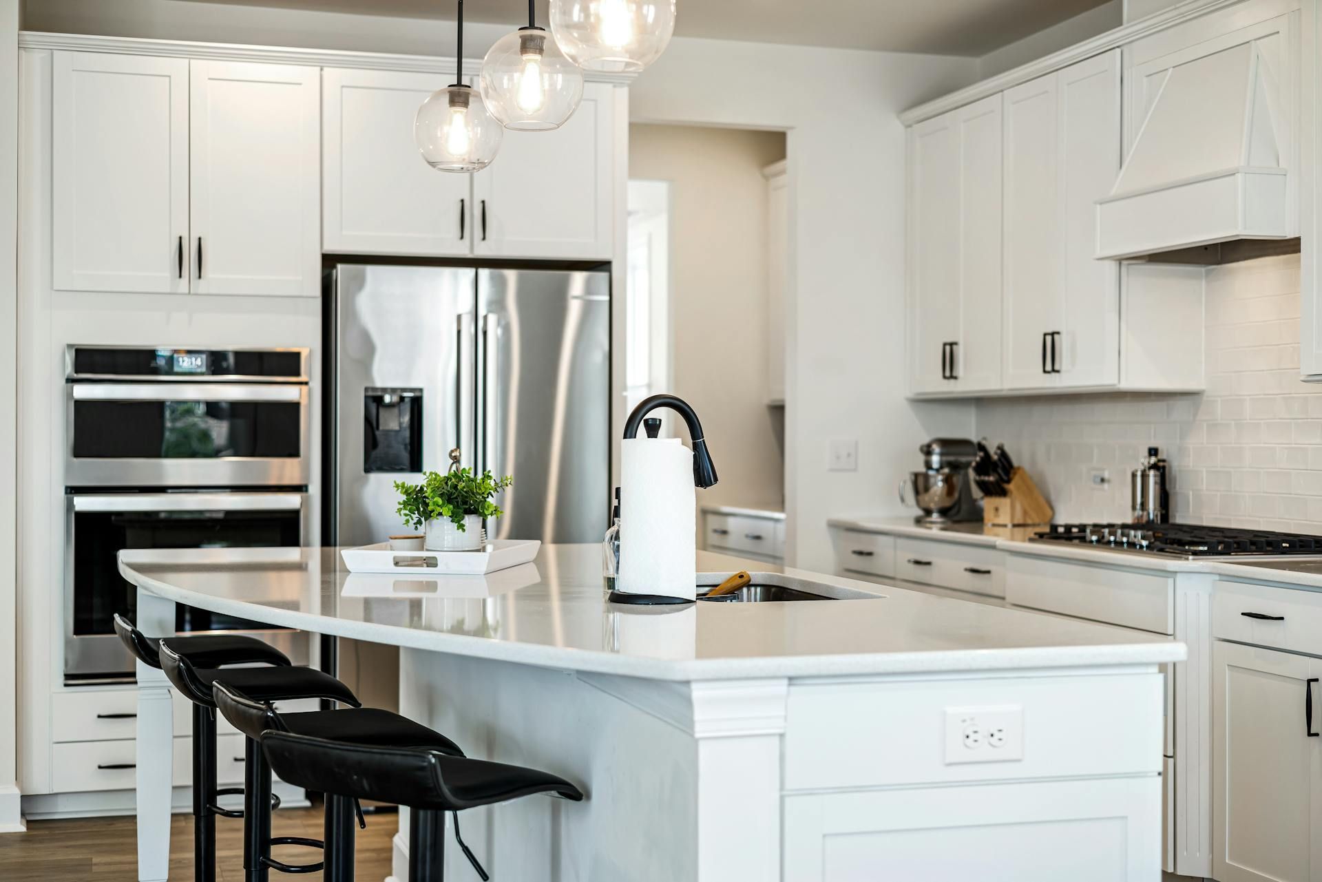 Bright white modern kitchen with island, black stools, stainless steel appliances, and pendant lights