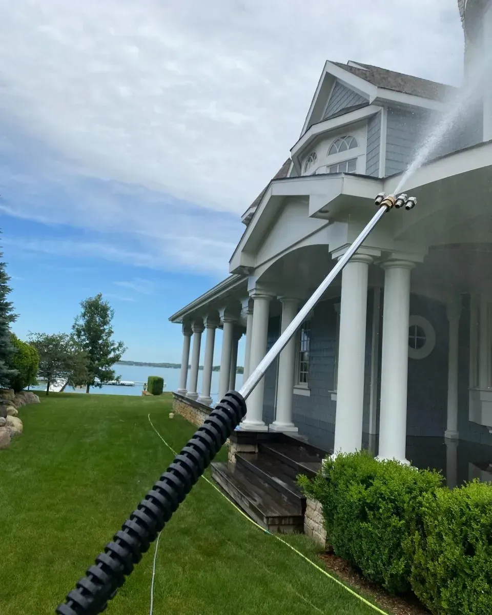 Person pressure-washing a gray house exterior beside a lawn and lake under a cloudy sky
