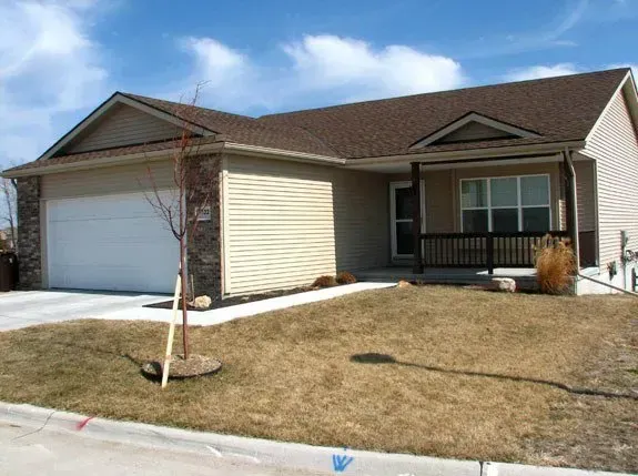 Single-story suburban house with attached garage, brown roof, small front lawn, and young tree.