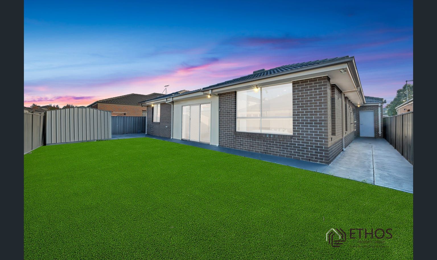 Exterior of a modern single-story house with dark brick siding, a small lawn, and a fenced backyard at dusk.