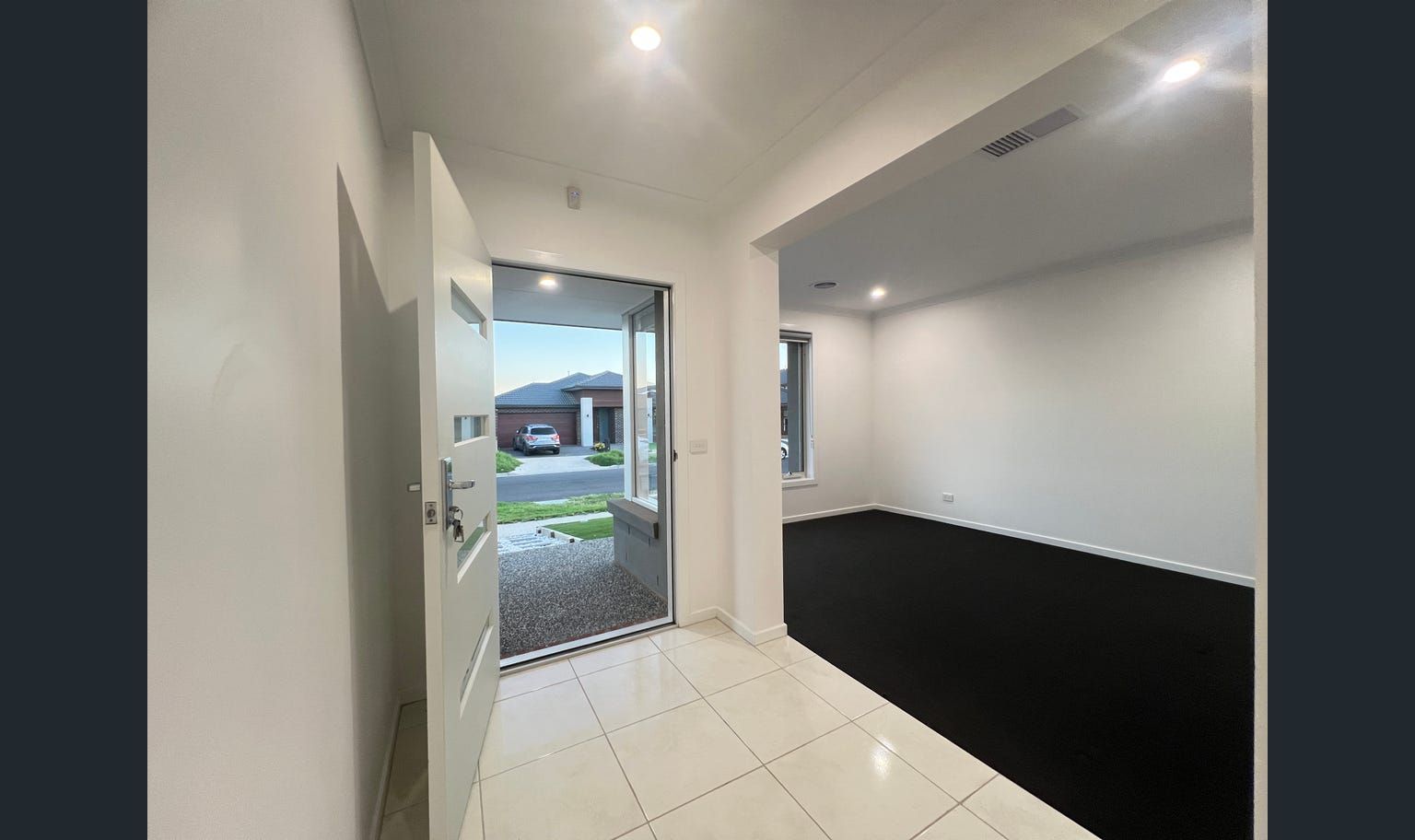 An entryway of a modern home. The door is open, with white walls and a dark carpeted living area visible.