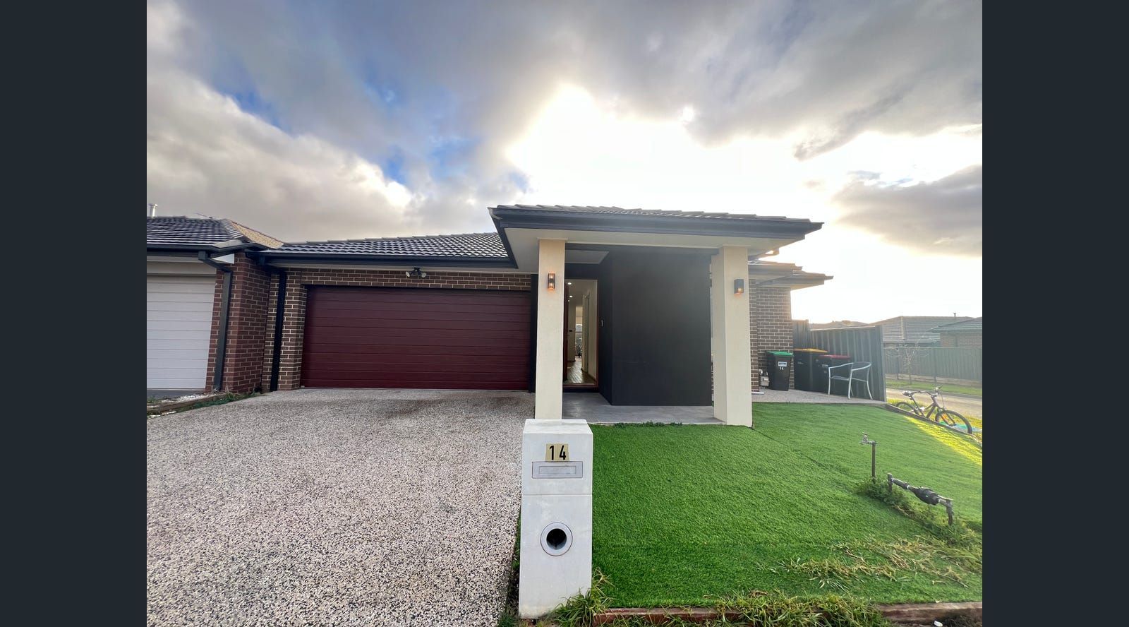 Modern house with a maroon garage door, green lawn, and gravel driveway under a cloudy sky.
