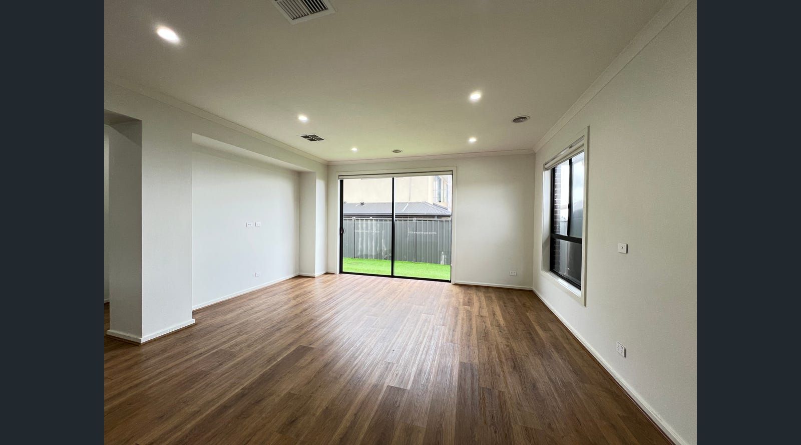 Empty living room with hardwood floors, white walls, and a sliding glass door leading to a fenced yard.