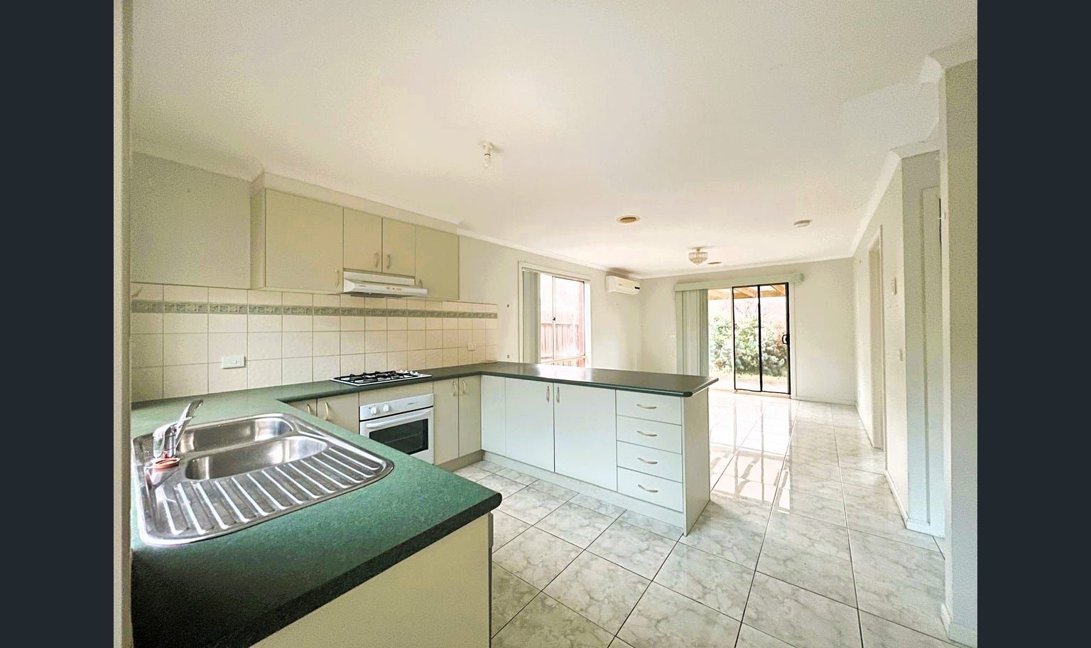 Empty kitchen with light-green countertops, white cabinets, and an island. The room opens to a sunlit living area.